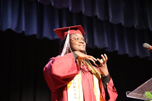 A graduate from the Alabama School for the Blind signs while on stage at graduation. She wears a maroon cap and gown and a yellow stole, indicating that she is valedictorian. 
